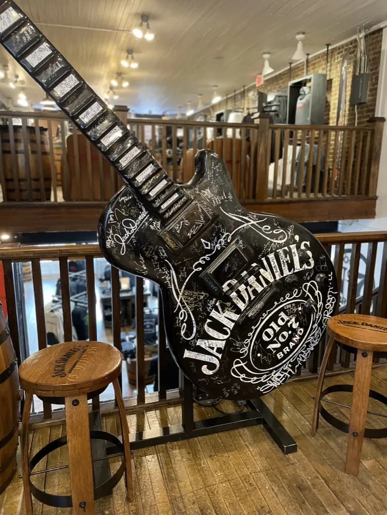 Oversized black Jack Daniel's Old No. 7 guitar covered in autographs, flanked by barrel stools, inside a Lynchburg shop