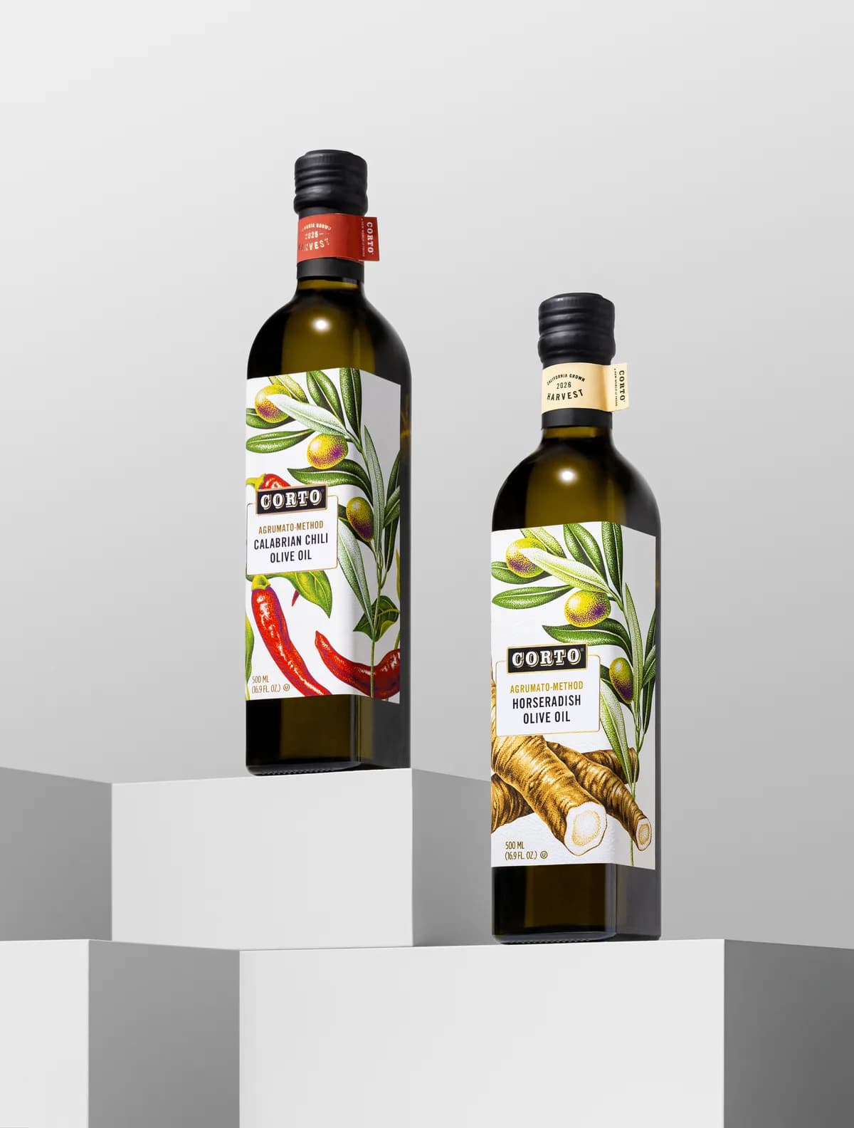 Two Corto Agrumato-Method olive oil bottles, Calabrian Chili and Horseradish, on white pedestals